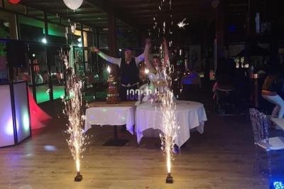 Des étincelles jaillissent de dispositifs au sol dans une salle décorée, avec des tables et des ballons suspendus.