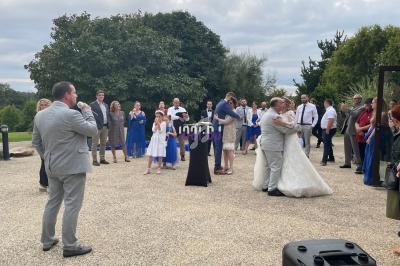 Un groupe de personnes assiste à une cérémonie en extérieur, avec un couple dansant au centre et un homme parlant au micro.