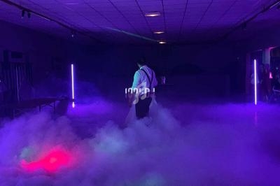 Un homme marche dans une salle sombre éclairée par des lumières violettes et entourée de fumée au sol.