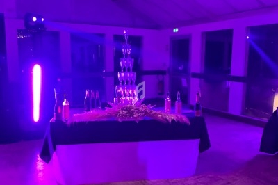 Tour de coupes en verre empilées sur une table décorée, entourée de bouteilles, éclairée par une lumière violette.