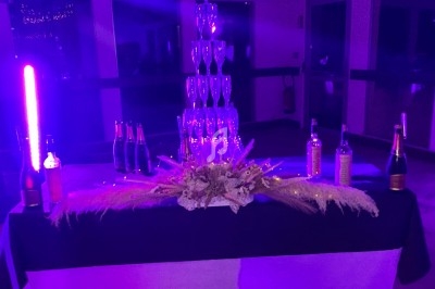 Tour de coupes en verre empilées sur une table décorée, entourée de bouteilles, éclairée par une lumière violette.