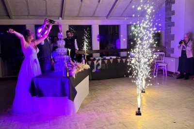Une femme en robe blanche lève les bras près d'une fontaine de champagne et de feux d'artifice en intérieur.