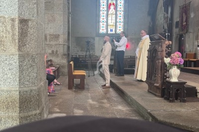 Un homme en costume est baptisé par un prêtre dans une église avec un vitrail coloré en arrière-plan.