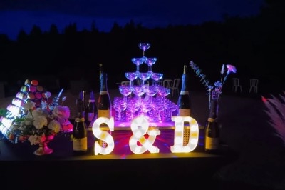 Tour de coupes de champagne illuminée entourée de bouteilles, fleurs et lettres décoratives ’S & D’ dans un décor nocturne.