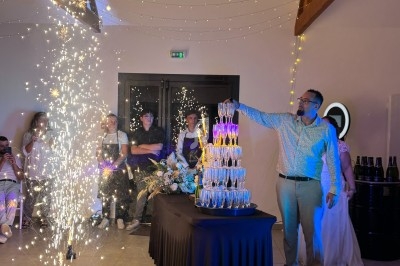 Un homme verse du champagne sur une pyramide de verres, entouré de convives et de décorations lumineuses.