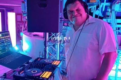 Un DJ souriant mixe de la musique sur une console, entouré d'éclairages colorés et d'équipements électroniques.