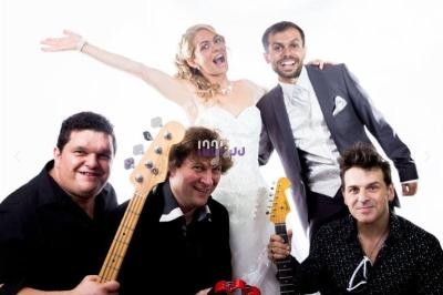 Un groupe de musiciens souriants pose avec une mariée en robe blanche et un homme en costume sur fond blanc.