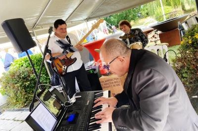 Musiciens jouant en plein air sous une tente, avec un pianiste au premier plan et un bassiste et un batteur en arrière-plan.