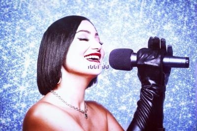 Femme souriante chantant dans un micro, portant des gants noirs et un collier, sur un fond scintillant bleu.