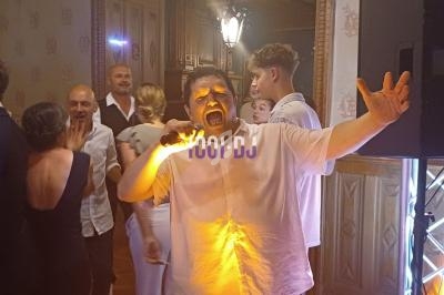 Un homme chante au micro dans une salle avec des invités en arrière-plan lors d'un événement festif.