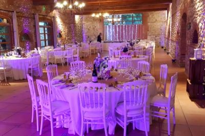 Salle de réception décorée pour un événement, avec tables rondes dressées, chaises blanches et éclairage tamisé.