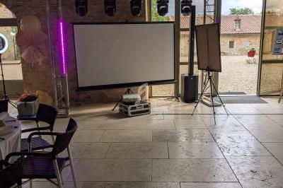 Salle en pierre avec un écran de projection, des projecteurs suspendus et des chaises disposées autour de tables.