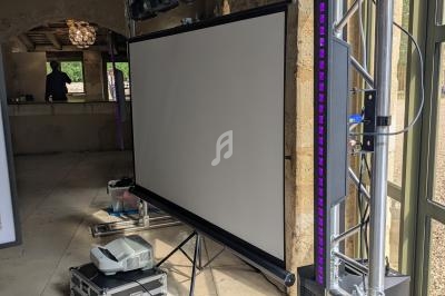 Écran de projection sur trépied entouré d'équipements audiovisuels dans une salle avec murs en pierre et poutres apparentes.