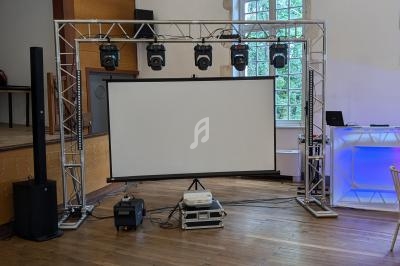 Structure métallique avec projecteurs, écran de projection et matériel audio dans une salle avec parquet et fenêtre en arc.