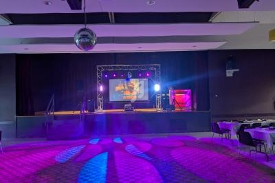 Salle de spectacle avec scène éclairée par des projecteurs colorés, écran central et tables disposées autour.