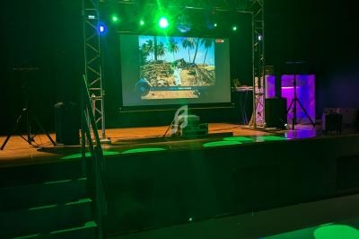 Scène éclairée avec projecteurs verts, écran affichant une image tropicale et équipements audio-visuels en arrière-plan.