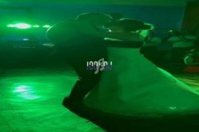 Un couple danse sous une lumière verte dans une salle sombre, entouré de spectateurs en arrière-plan.