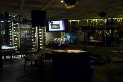 Salle sombre avec tables, chaises, écran de projection allumé et étagères de bouteilles éclairées en arrière-plan.