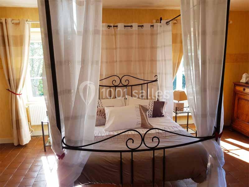 Lit à baldaquin avec voilages blancs dans une chambre lumineuse aux murs jaunes, sol en terre cuite et mobilier en bois.