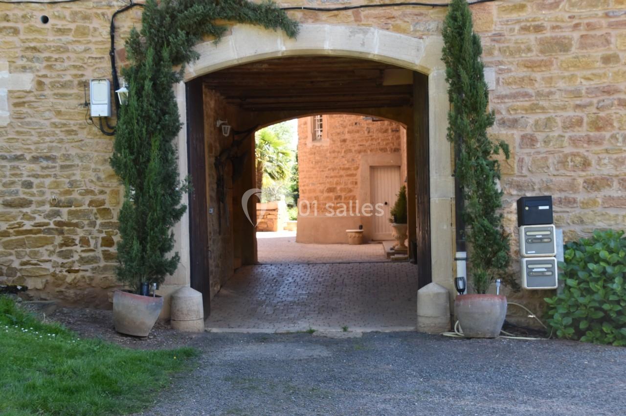 Passage voûté en pierre avec des plantes en pot, menant à une cour intérieure pavée et lumineuse.