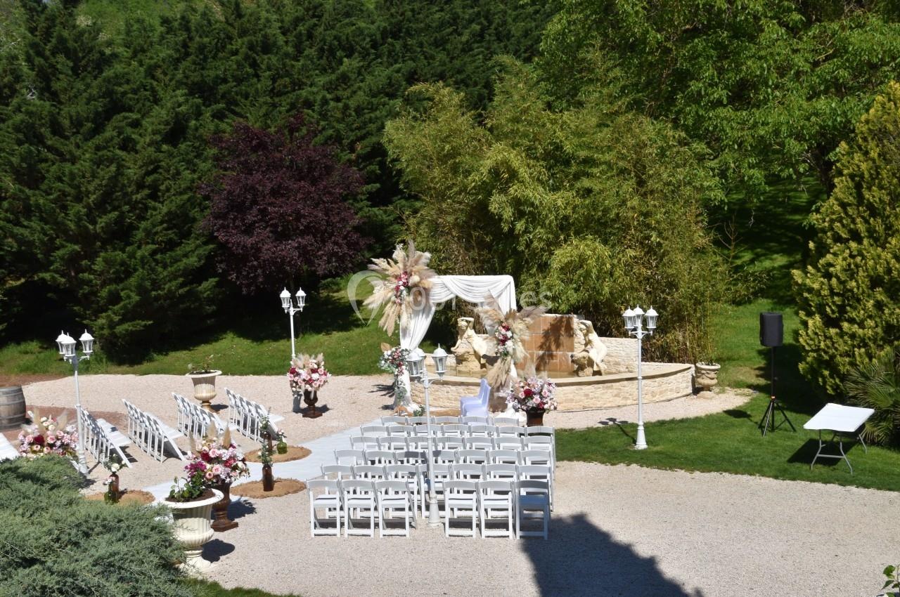 Chaises blanches alignées devant une arche décorée de fleurs et de drapés, dans un jardin verdoyant.