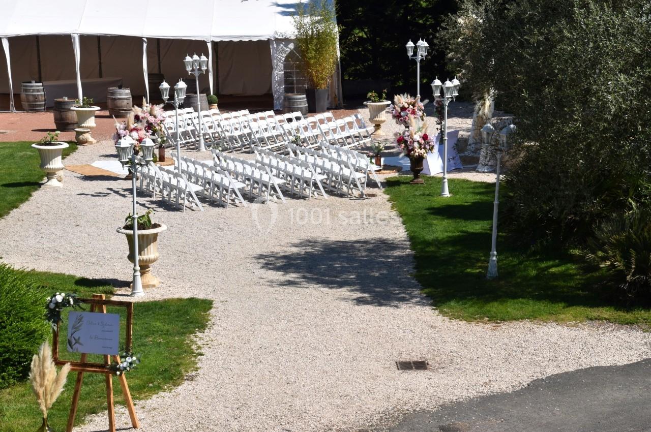 Allée gravillonnée menant à une cérémonie en plein air avec chaises blanches, décorations florales et tente blanche.