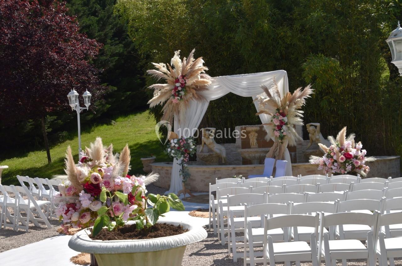 Cérémonie en plein air avec arche décorée de fleurs et de pampas, entourée de chaises blanches alignées.