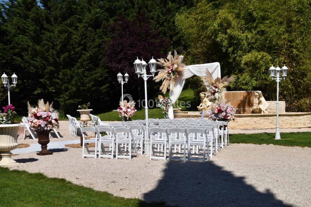 Chaises blanches alignées devant une arche décorée de fleurs pour une cérémonie en extérieur, entourée de verdure.
