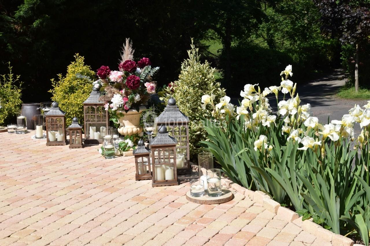 Lanternes décoratives entourées de fleurs blanches et rouges sur une terrasse pavée, bordée de verdure.