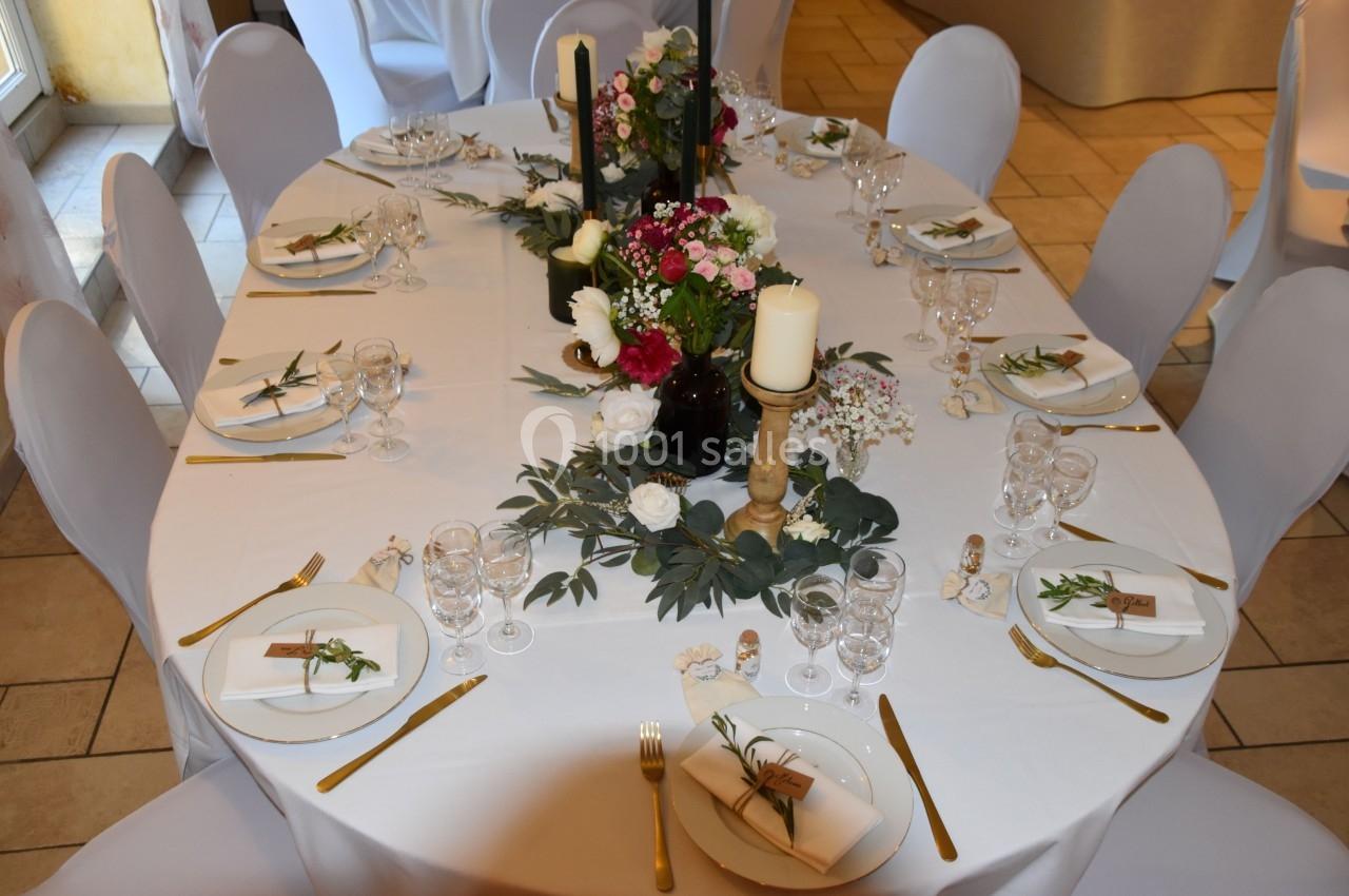 Table décorée pour un événement, avec nappes blanches, bougies, fleurs et couverts dorés disposés élégamment.