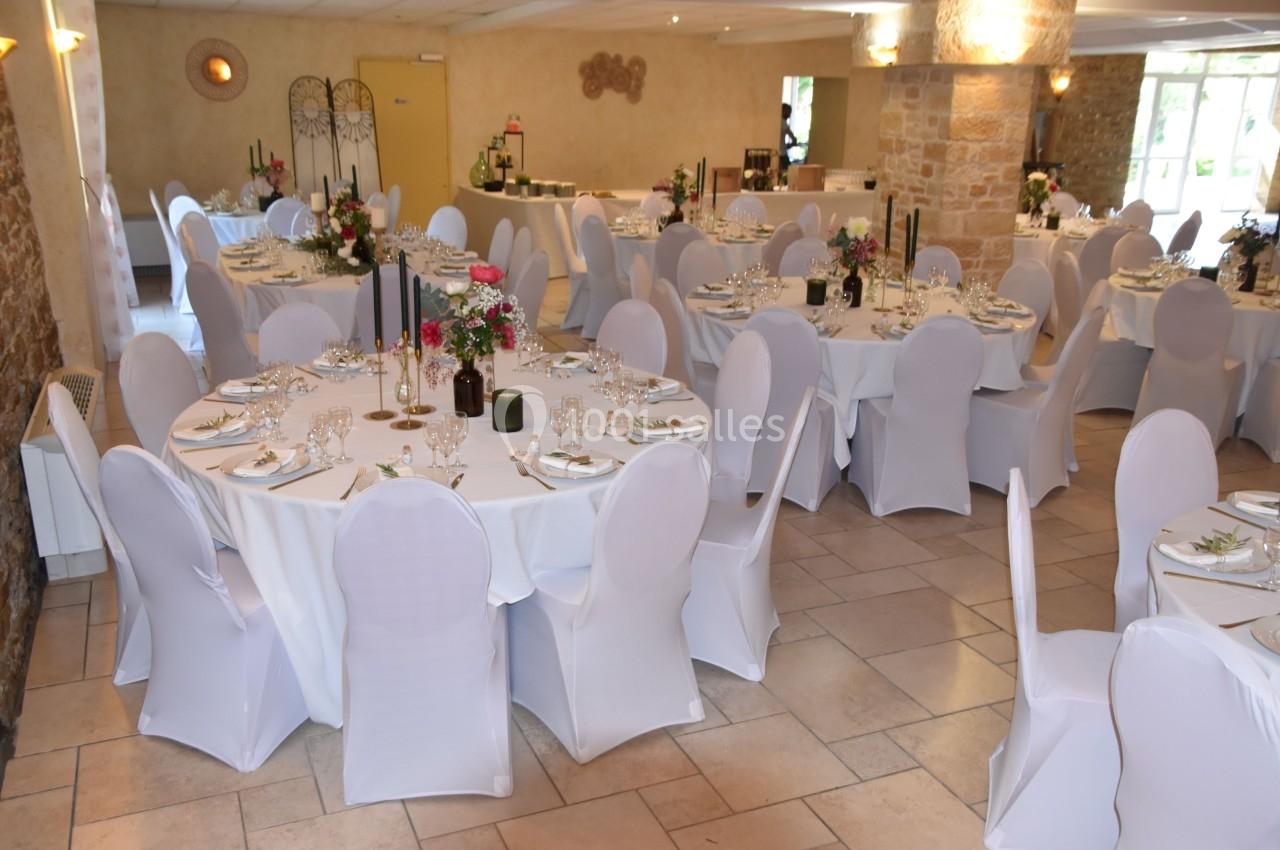Salle de réception décorée avec des tables rondes, nappes blanches, chaises houssées et centres de table fleuris.