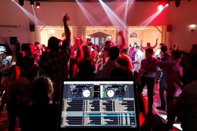 Vue d'une fête en intérieur avec des personnes dansant sous des lumières colorées, écran de DJ au premier plan.