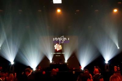 Salle sombre avec un DJ sur scène, éclairée par des faisceaux lumineux et entourée d'un public en soirée.