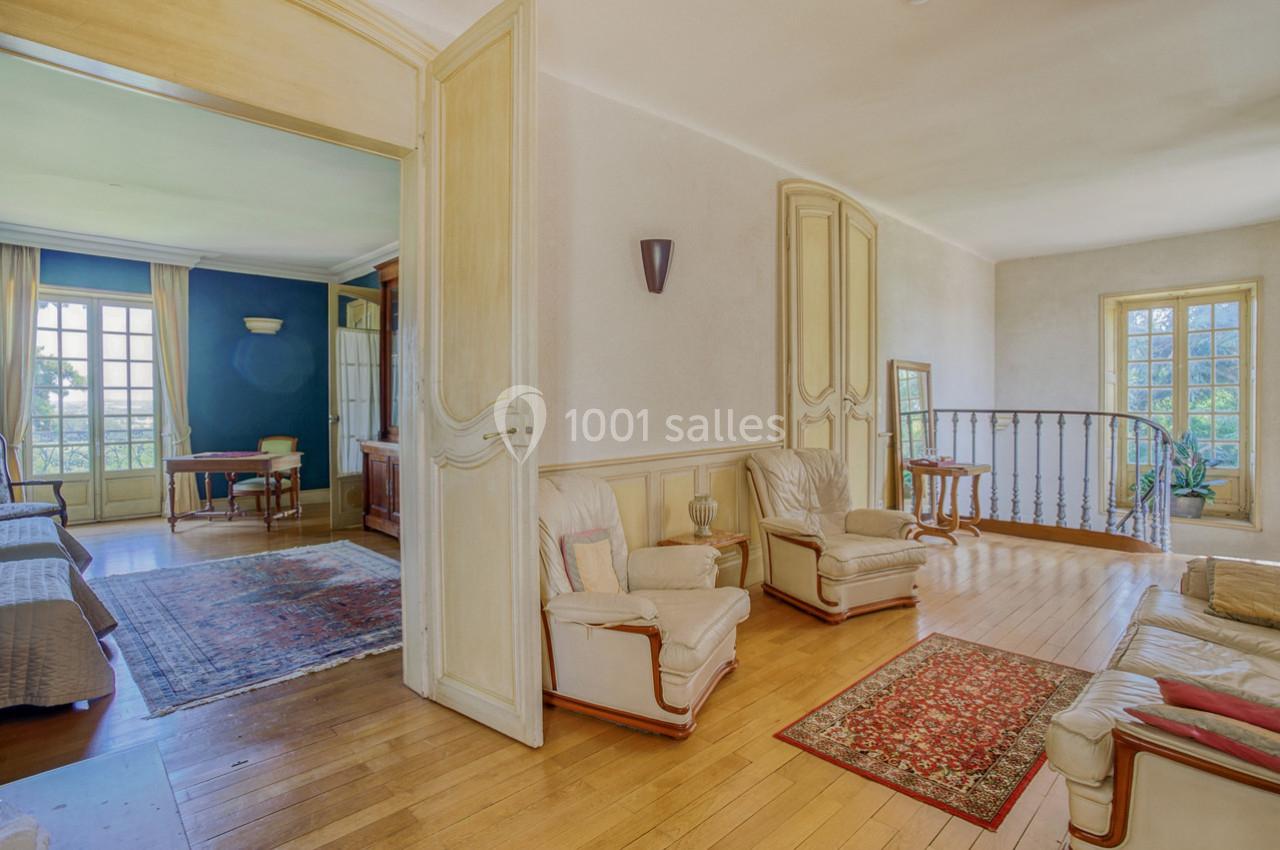 Salon lumineux avec parquet, fauteuils en cuir, tapis colorés, ouvert sur une pièce adjacente et un escalier en fer forgé.