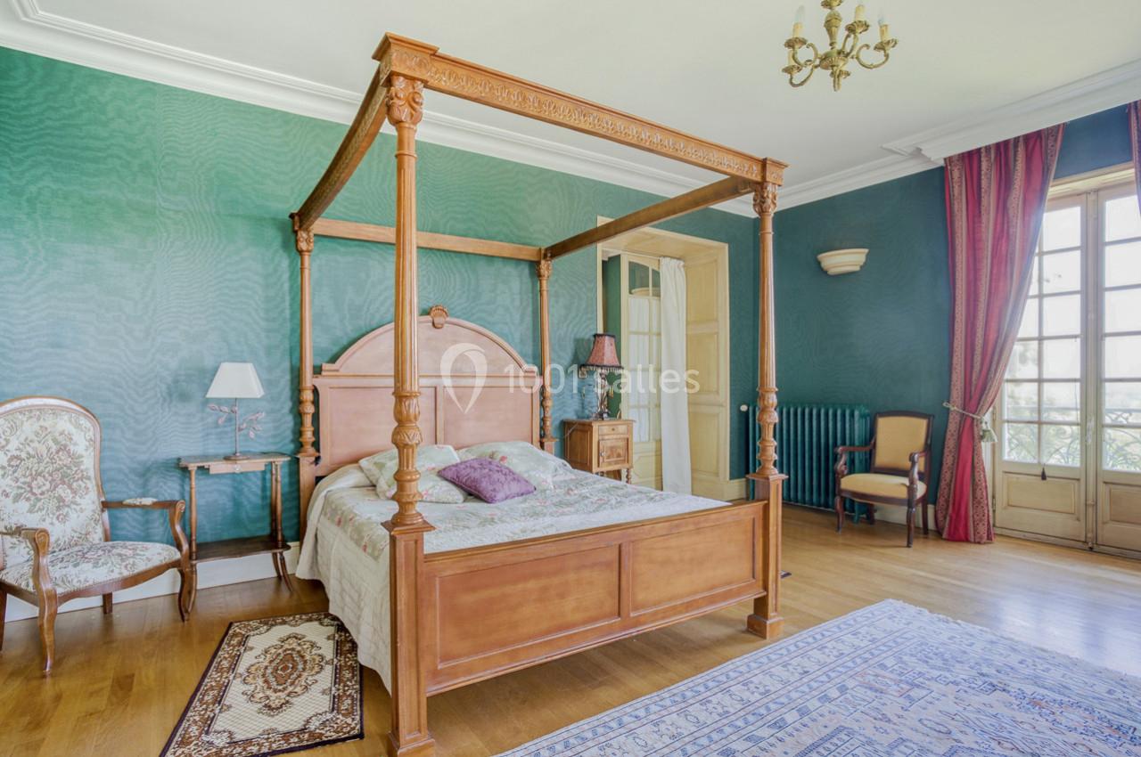 Chambre avec lit à baldaquin en bois, murs verts, parquet, tapis, fauteuils et grande fenêtre avec rideaux rouges.