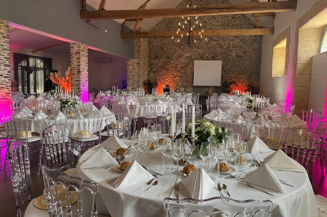 Salle de réception décorée avec des tables rondes élégantes, nappes blanches et chaises transparentes sous des poutres…