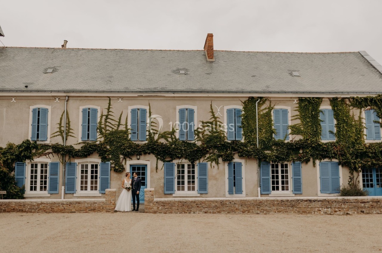 Façade d'une grande maison ancienne avec volets bleus, recouverte partiellement de lierre, un couple debout devant.