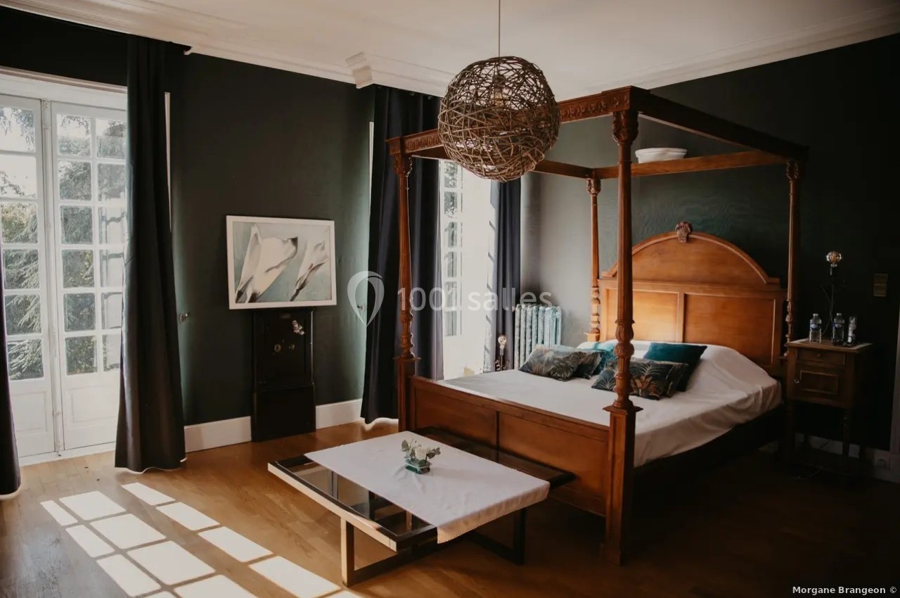 Chambre lumineuse avec lit à baldaquin en bois, table basse blanche et murs sombres, éclairée par une grande fenêtre.