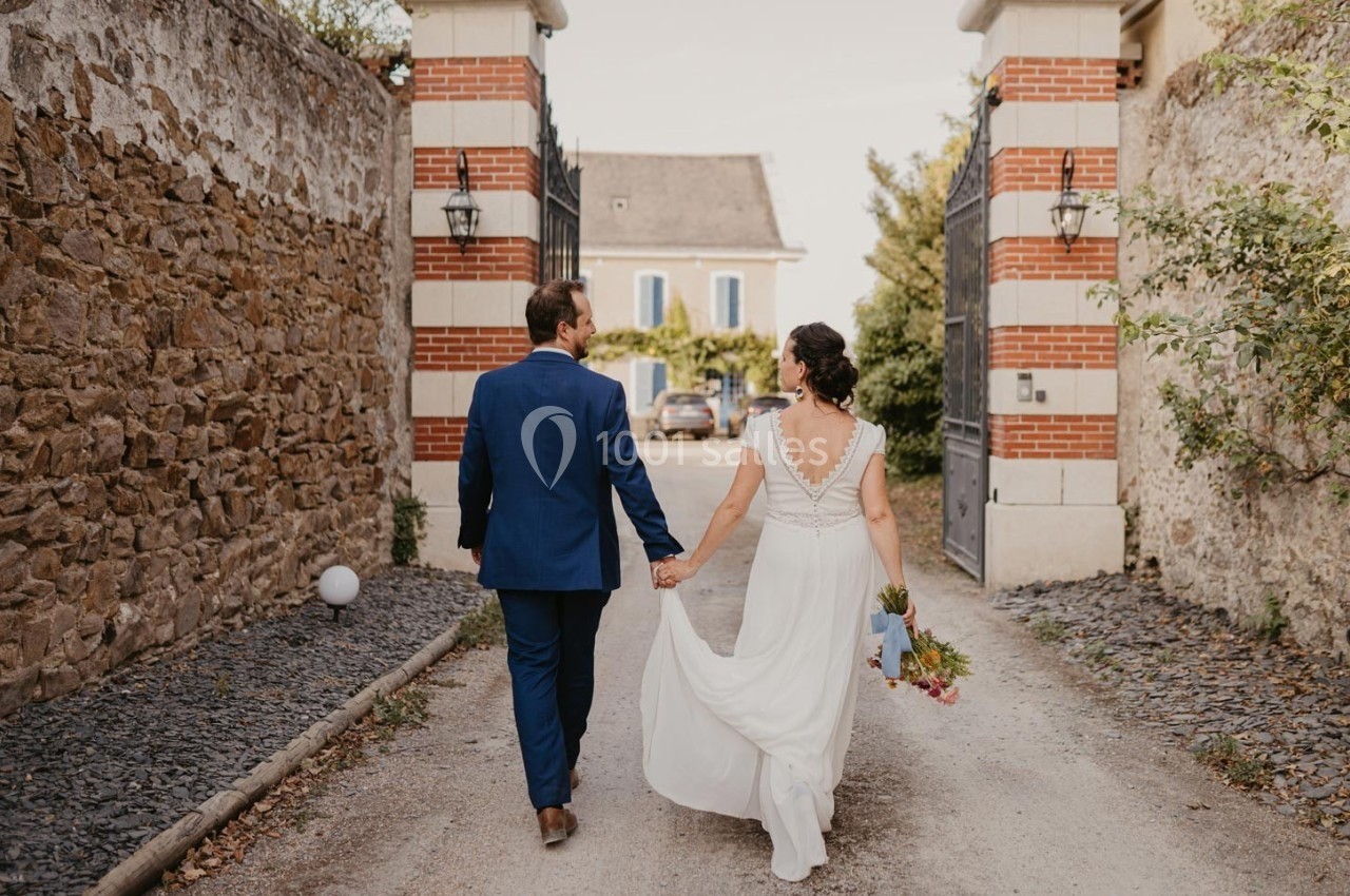 Un couple en tenue de mariage marche main dans la main sur une allée bordée de murs en pierre et en briques.