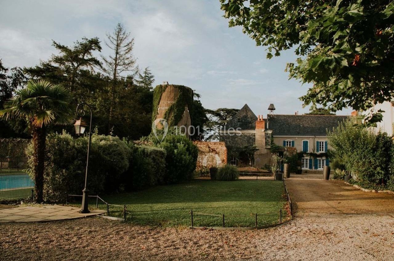 Jardin avec pelouse, arbres, piscine, et bâtiment ancien en pierre avec volets bleus sous un ciel partiellement nuageux.