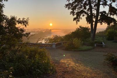 Lever de soleil illuminant un jardin avec des arbres, un muret en pierre et une vue sur un paysage brumeux.