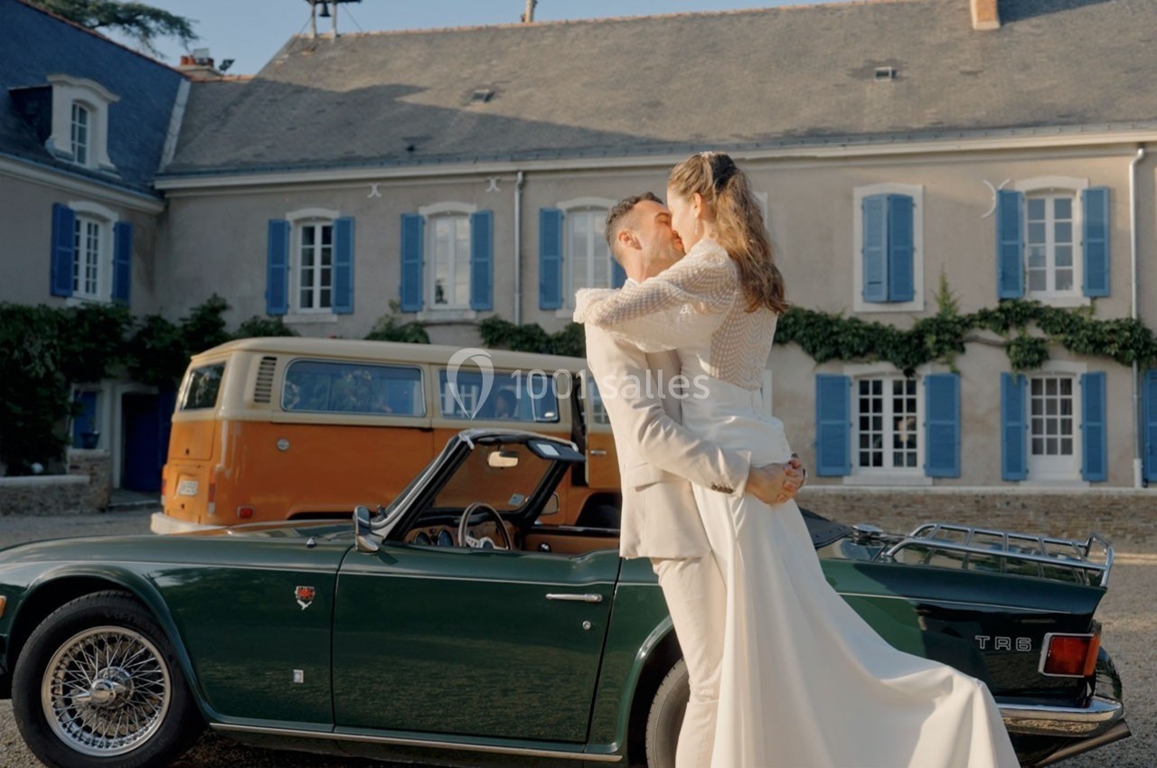 Un couple en tenue de mariage s'enlace devant une voiture ancienne verte et un van orange, près d'une maison.