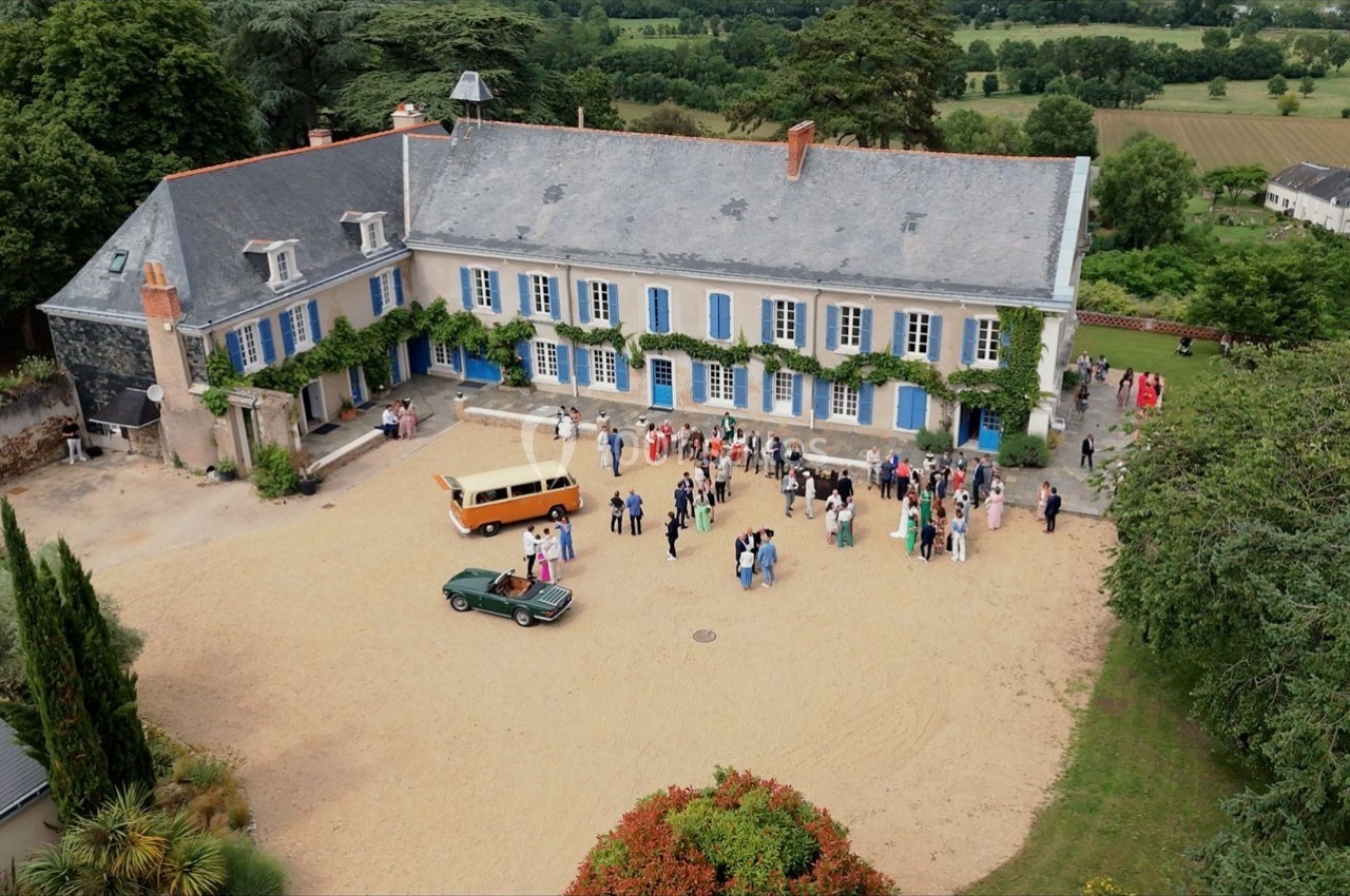 Vue aérienne d'une grande maison avec des volets bleus, entourée de verdure, et des personnes rassemblées dans la cour.