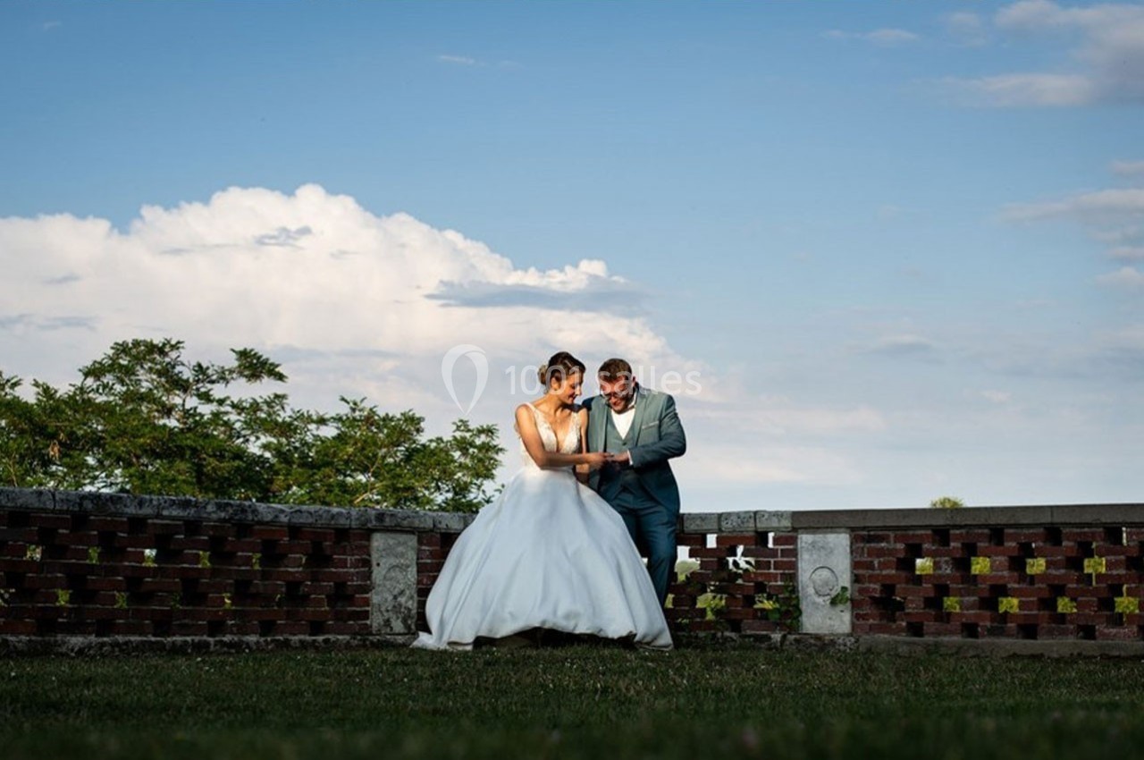 Un couple en tenue de mariage se tient devant un muret en briques, sous un ciel partiellement nuageux.
