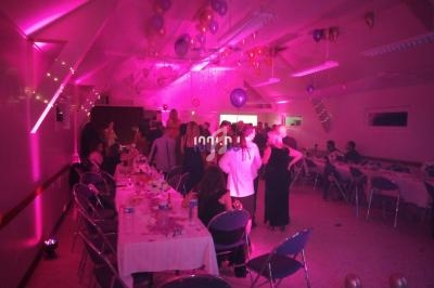 Salle décorée avec des ballons et éclairée en rose, des invités debout discutent près de tables dressées.