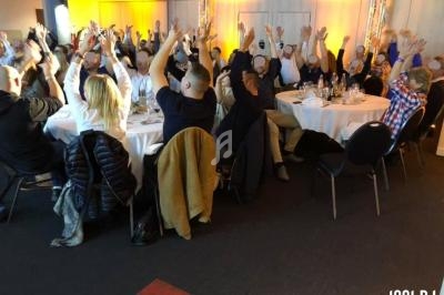Des personnes assises autour de tables rondes lèvent les bras dans une salle éclairée par des lumières chaudes.