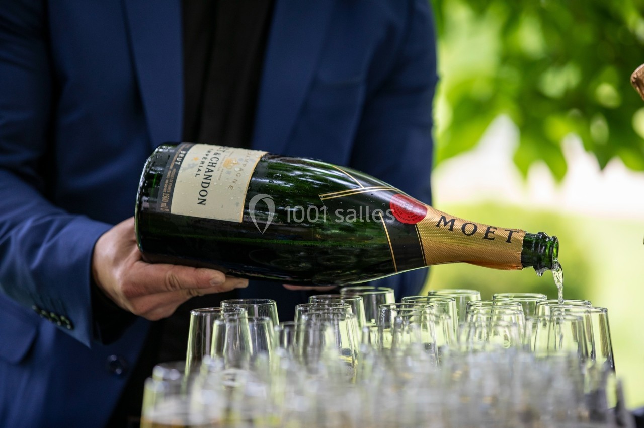 Une personne verse du champagne dans des flûtes alignées, en extérieur avec un fond de verdure.