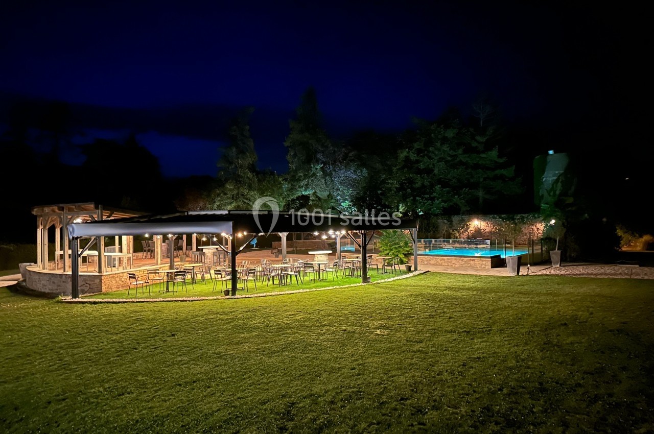 Espace extérieur éclairé de nuit avec terrasse couverte, tables, chaises et piscine entourée de verdure.