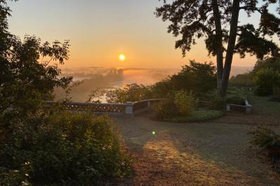 Lever de soleil sur un jardin avec des arbres, un chemin en terre et une balustrade en pierre surplombant un paysage brumeux.
