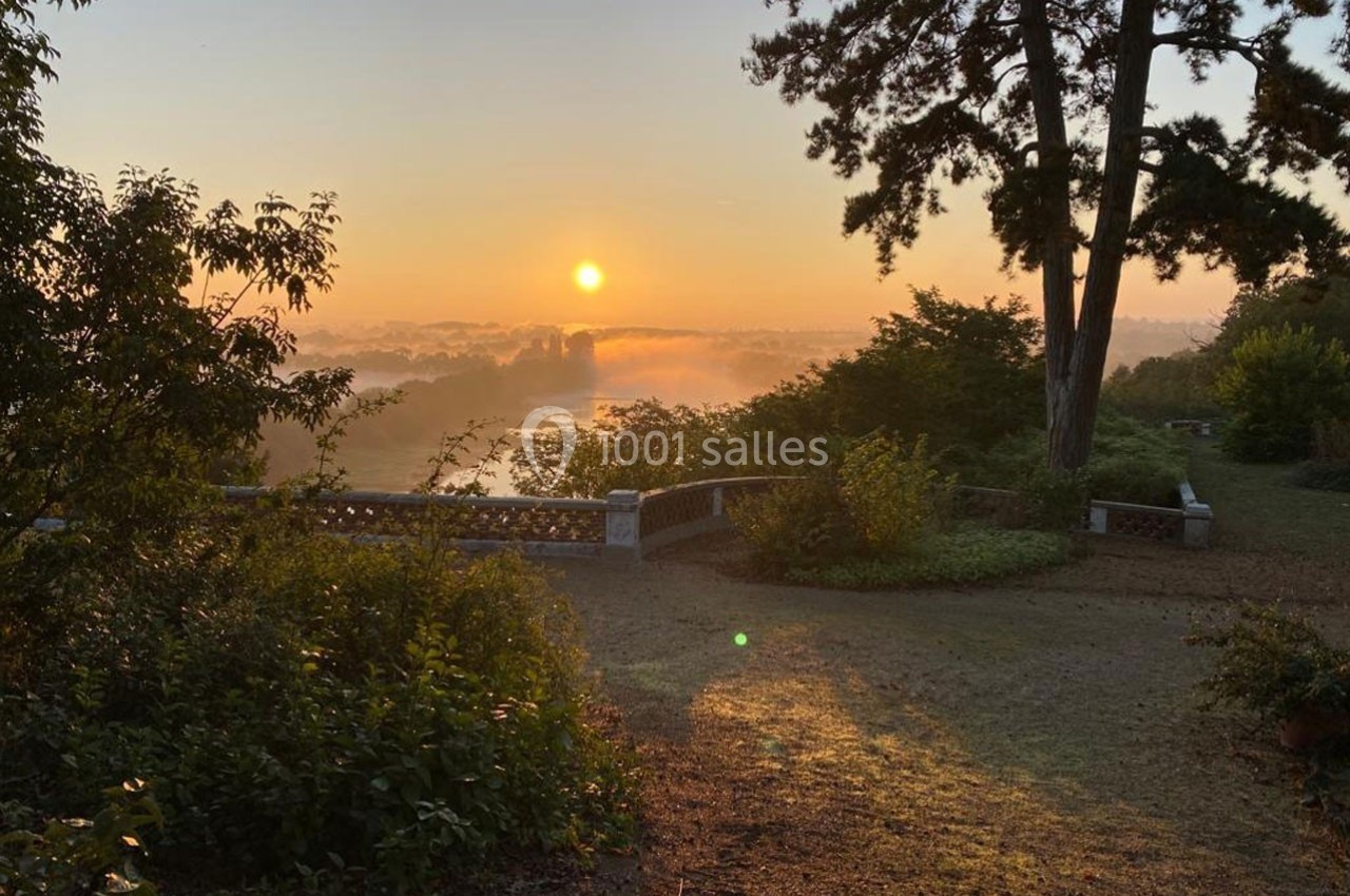 Lever de soleil sur un jardin avec des arbres, un chemin en terre et une balustrade en pierre surplombant un paysage brumeux.